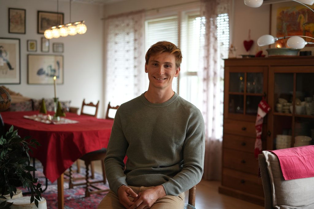 En ung mann med rødbrunt hår sitter i en spisestue og smiler.