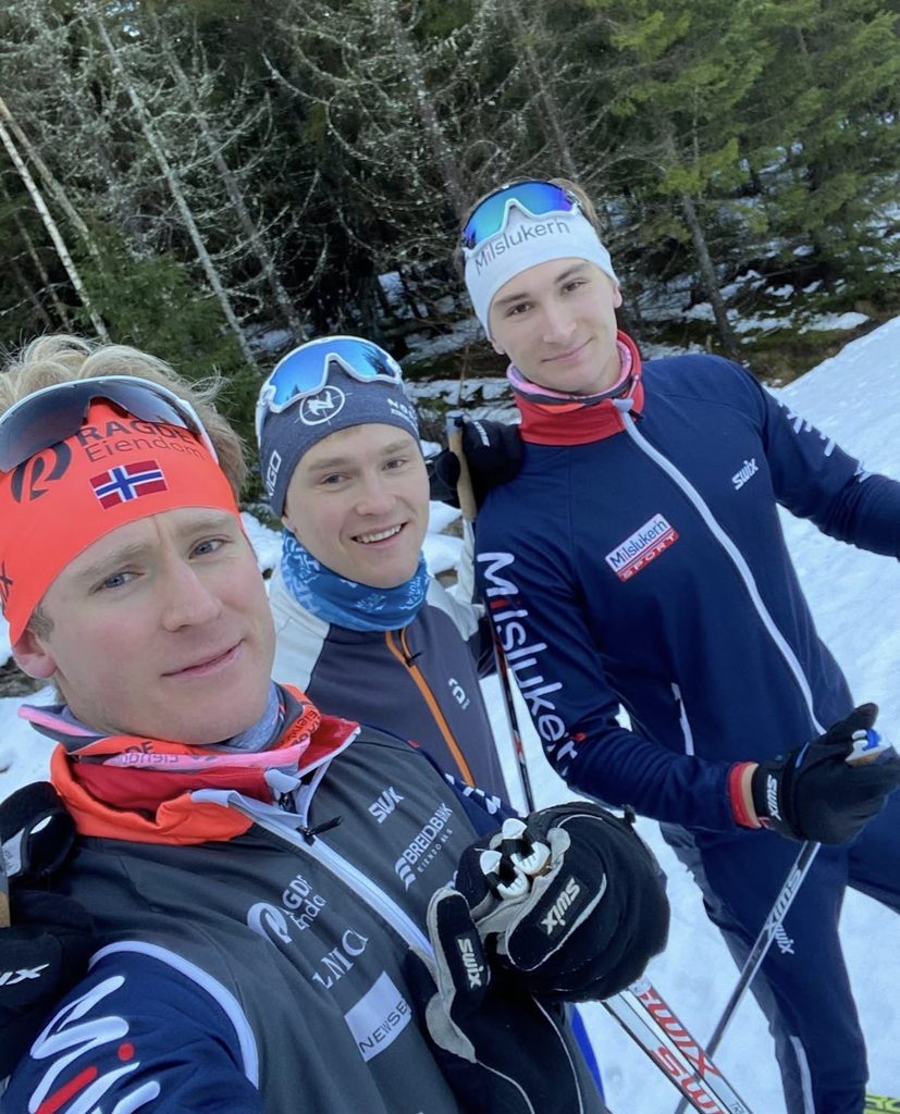 Tre langrennsløpere i treningsklær og med pannebånd som poserer for en selfie på en snødekt løype.