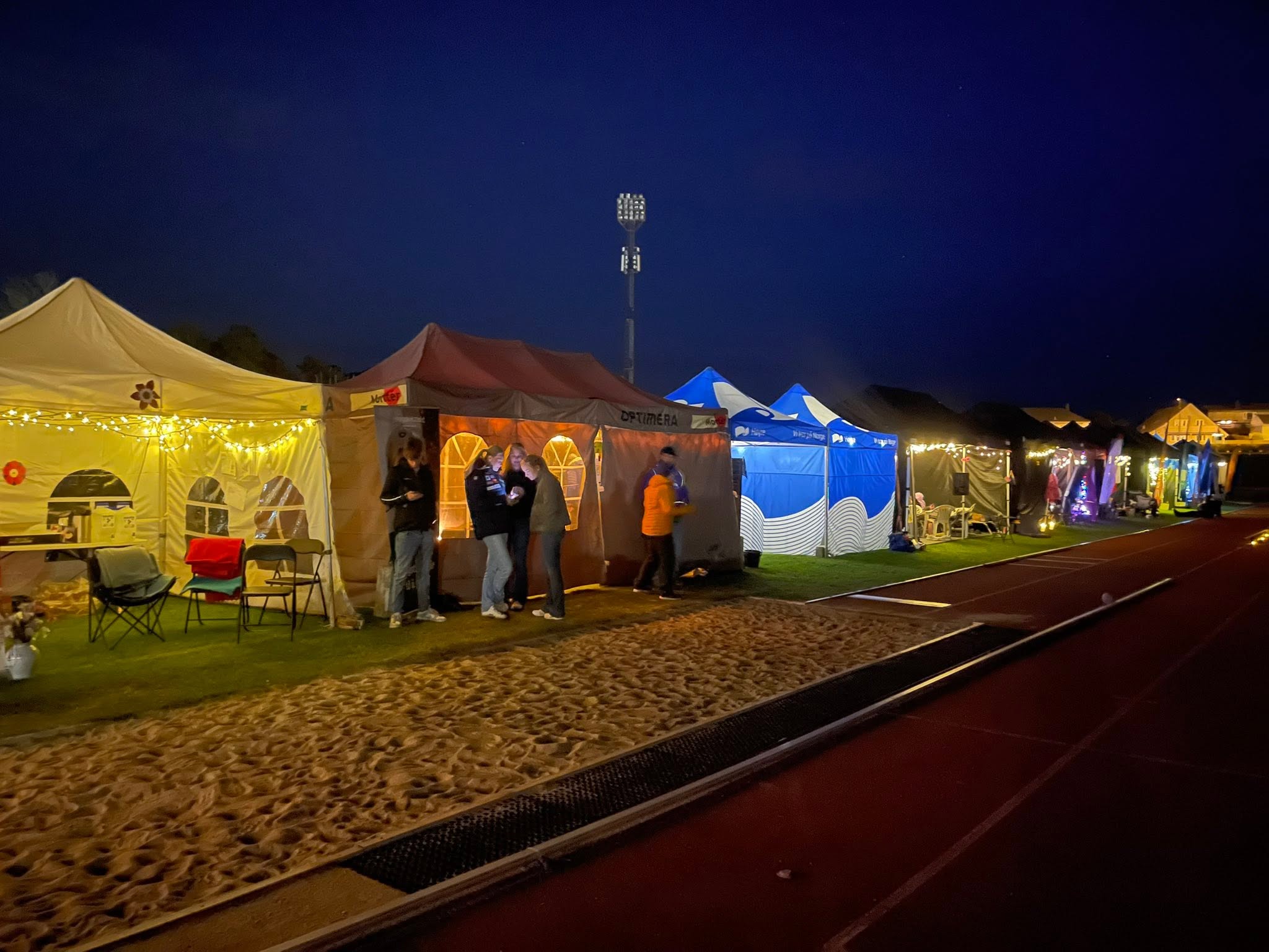 En rekke opplyste festtelt som er satt opp på en friidrettsbane om natten, med folk som står og snakker utenfor.
