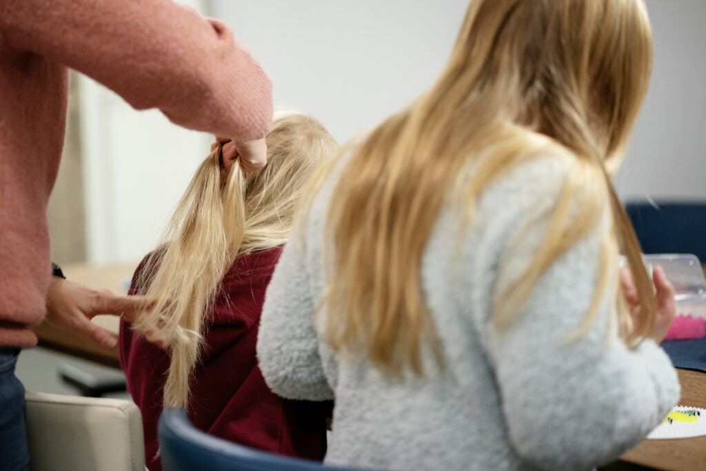 En person i en rosa genser fletter håret til en liten jente, mens et annet barn sitter i nærheten.