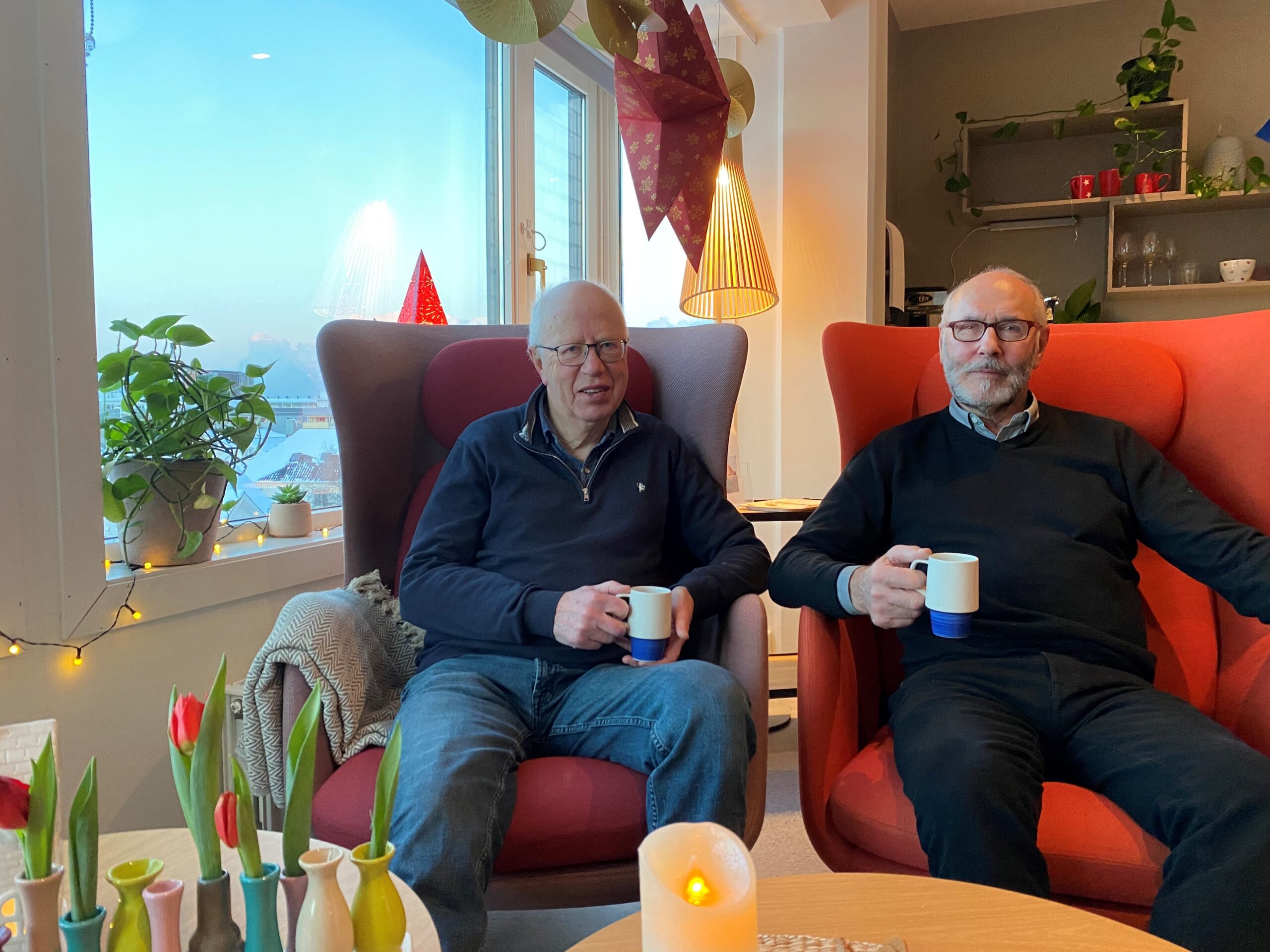 To menn som sitter i oransje lenestoler, hver med en kopp i hånden, i et rom med et stort vindu.