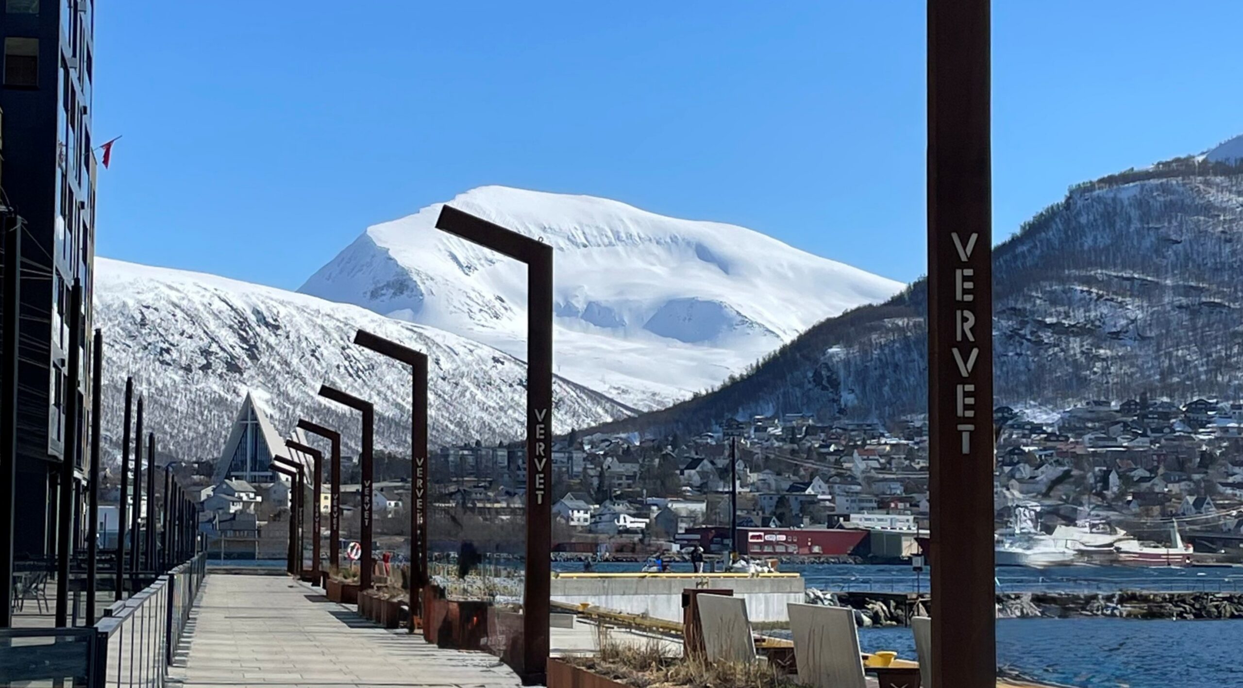 En gangvei langs vannkanten med skilt merket «VERVET», med utsikt over en havneby og snødekte fjell.